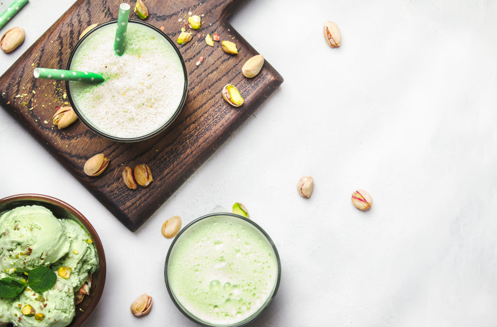 two bowls of Pistachio Smoothie, one was placed on top of a wood chopping board and a bowl of pistachio ice cream on the side of another bowl of pistachio smoothie with scattered pistachio on top of the table