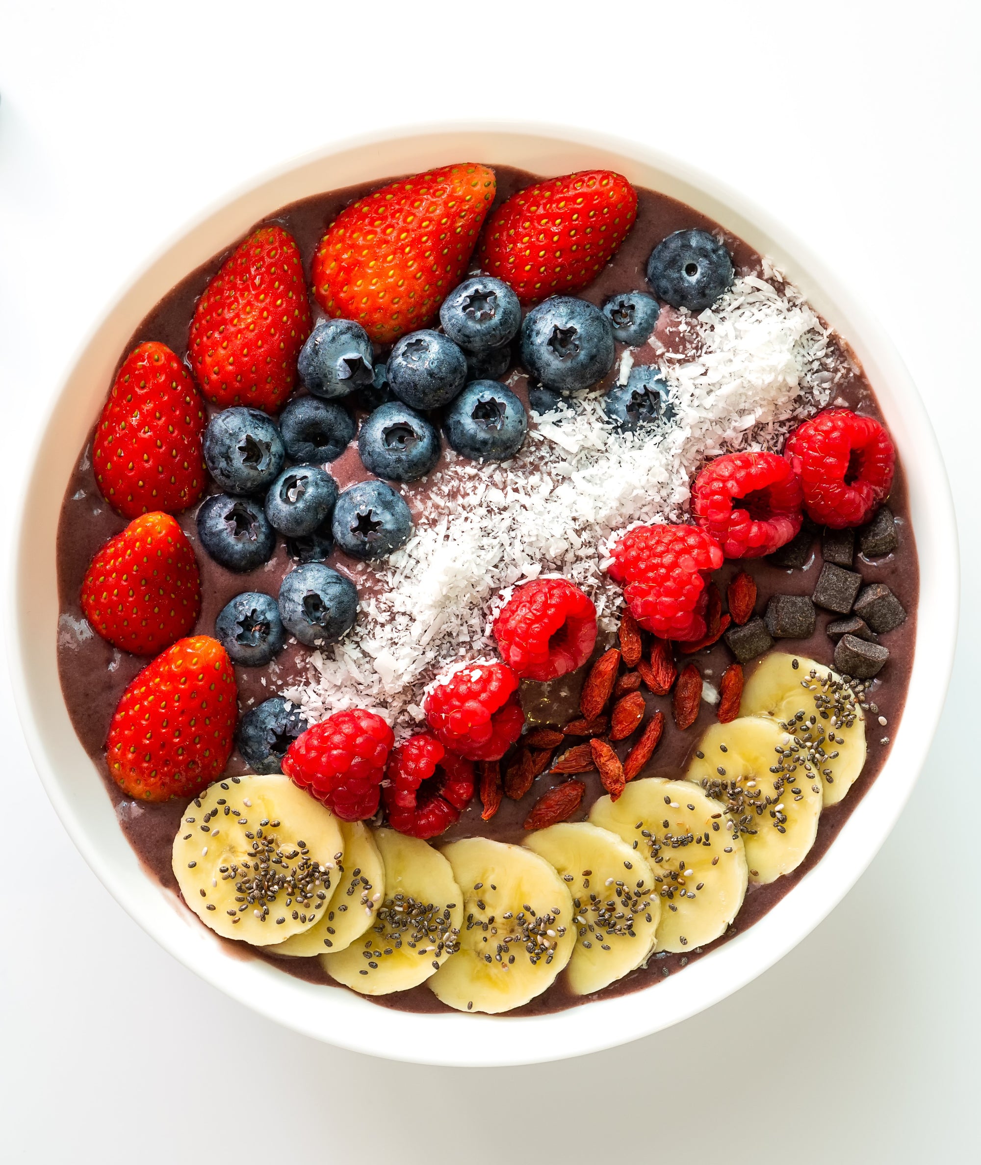 top view of a bowl of revive beets + roots acai bowl topped with slices of banana, chia seeds, berries, granola on top of a white table