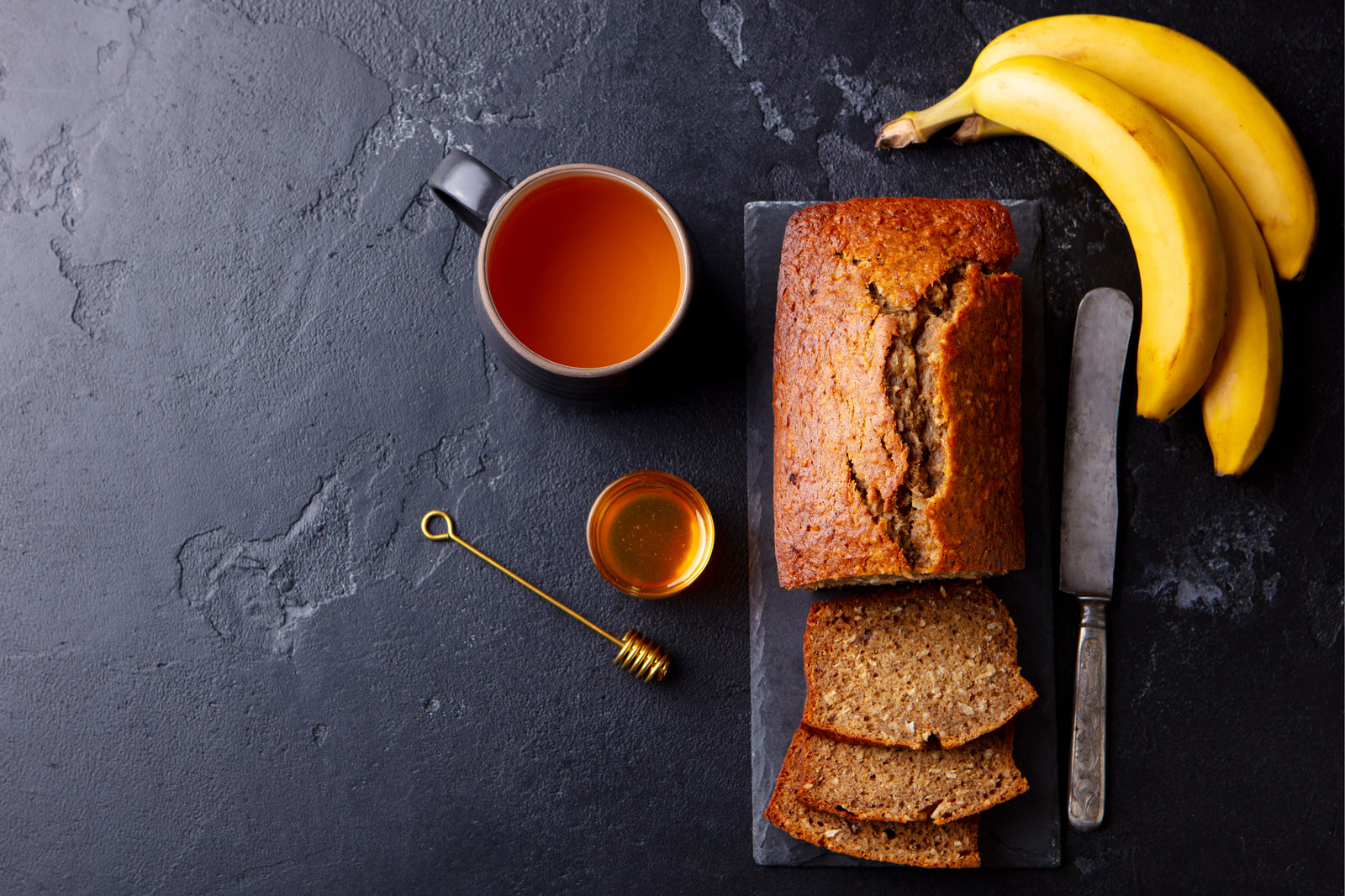 top view of Pineapple Chia Banana Bread on top of a concrete plank surrounded by bread-knife, three bananas, a cup of tea, a small cup of honey with honey dipper on top of a black-painted concrete table
