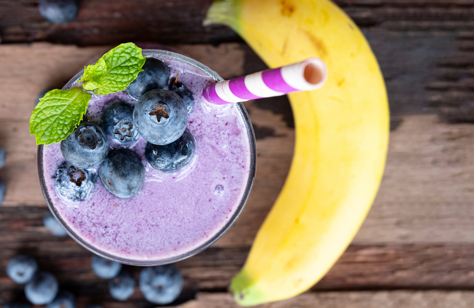 top view of a glass of pineapple chia cleanse topped with berries and mint with violet patterned paper straw and with berries and banana on the side on top of a wooden table