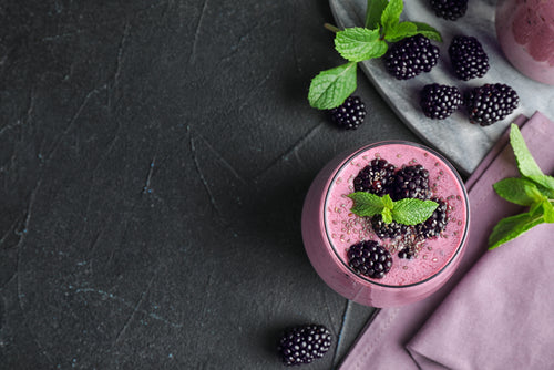 a glass of blackberry smoothie with blackberries toppings and mint leaves on top with blackberries and mint leaves side by side with pink folded table clothe