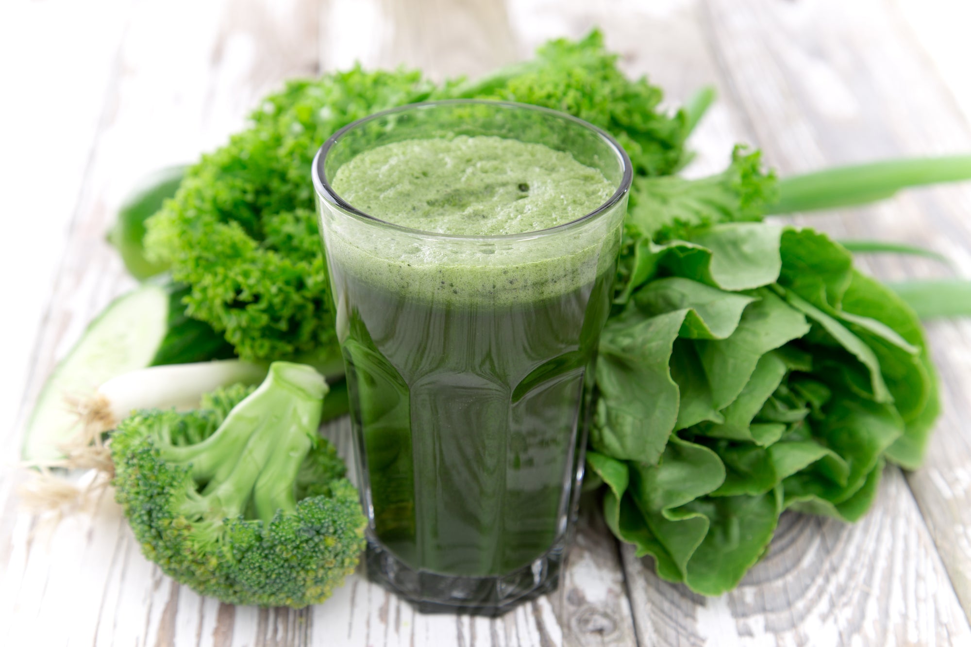 a glass of green smoothie surrounded by green vegetables on a wooden table.