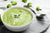 top view of a bowl with creamy broccoli soup with a spoon on the side on top of a circle-shaped wood with broccoli florets on top of a marble table