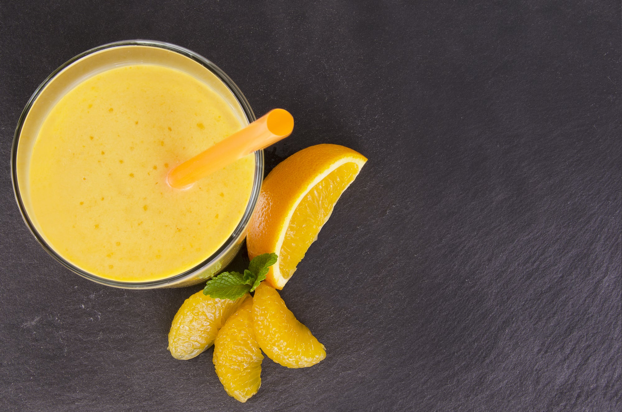 top view shot of a glass of Citrus Immunity smoothie with a yellow straw beside orange slices on a black surface.