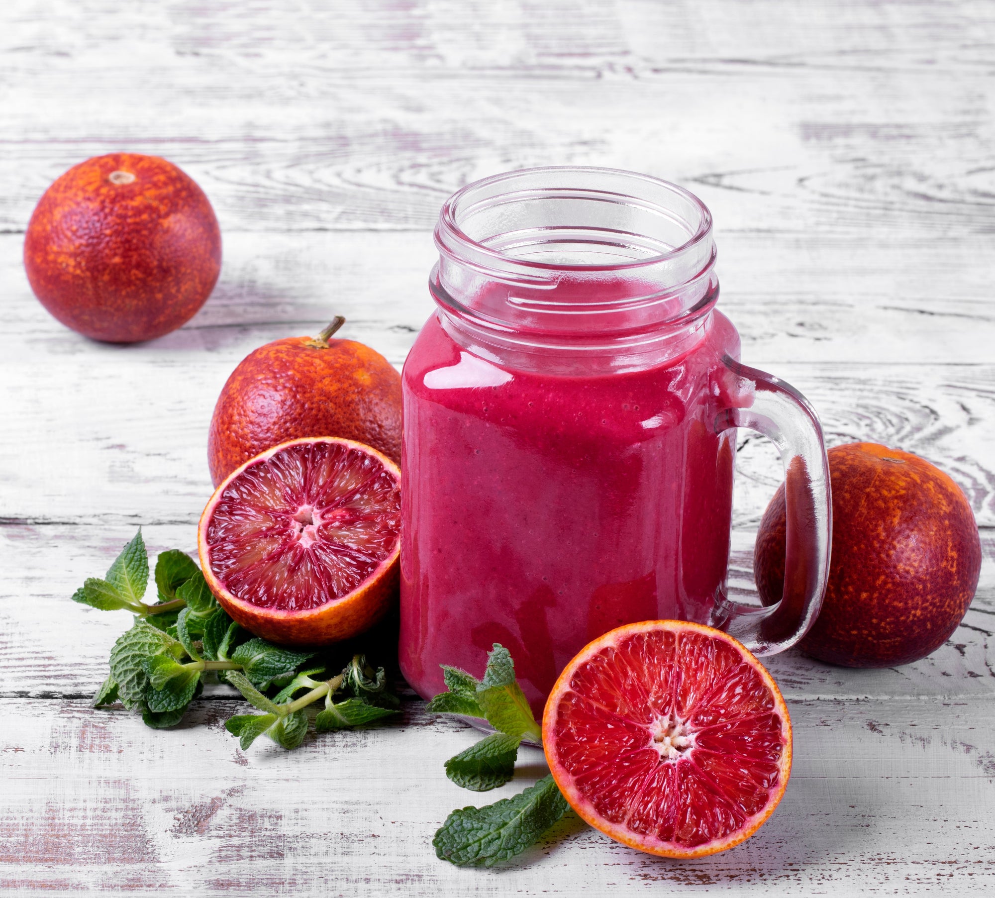 side view of a mason jar filled with blood orange greens smoothie surrounded by blood oranges,half-cut blood orange, and mint leaves on top of a wooden table