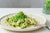 side view of a white ceramic plate with Pesto Zoodles with a fork on the side and a small jar of organic pressed greens on top of a white table