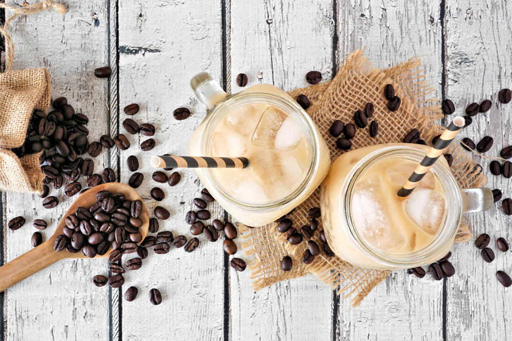 top view of two jars of iced smoothie coffee with black patterned paper straw on top of two pieces weaved cloth surrounded by coffee beans and a wooden spoon full of coffee beans