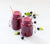 side view of two mason jar of berry coconut pineapple smoothie with light orange paper straw surrounded by blueberries and mint on top of a white table