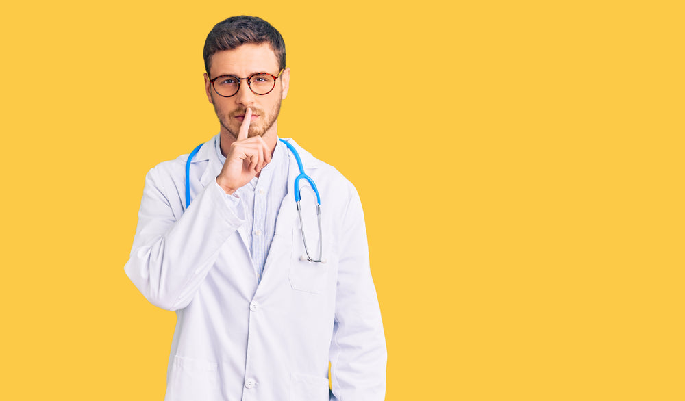 Image of a male doctor in a shushing hand gesture against a yellow wall. 
