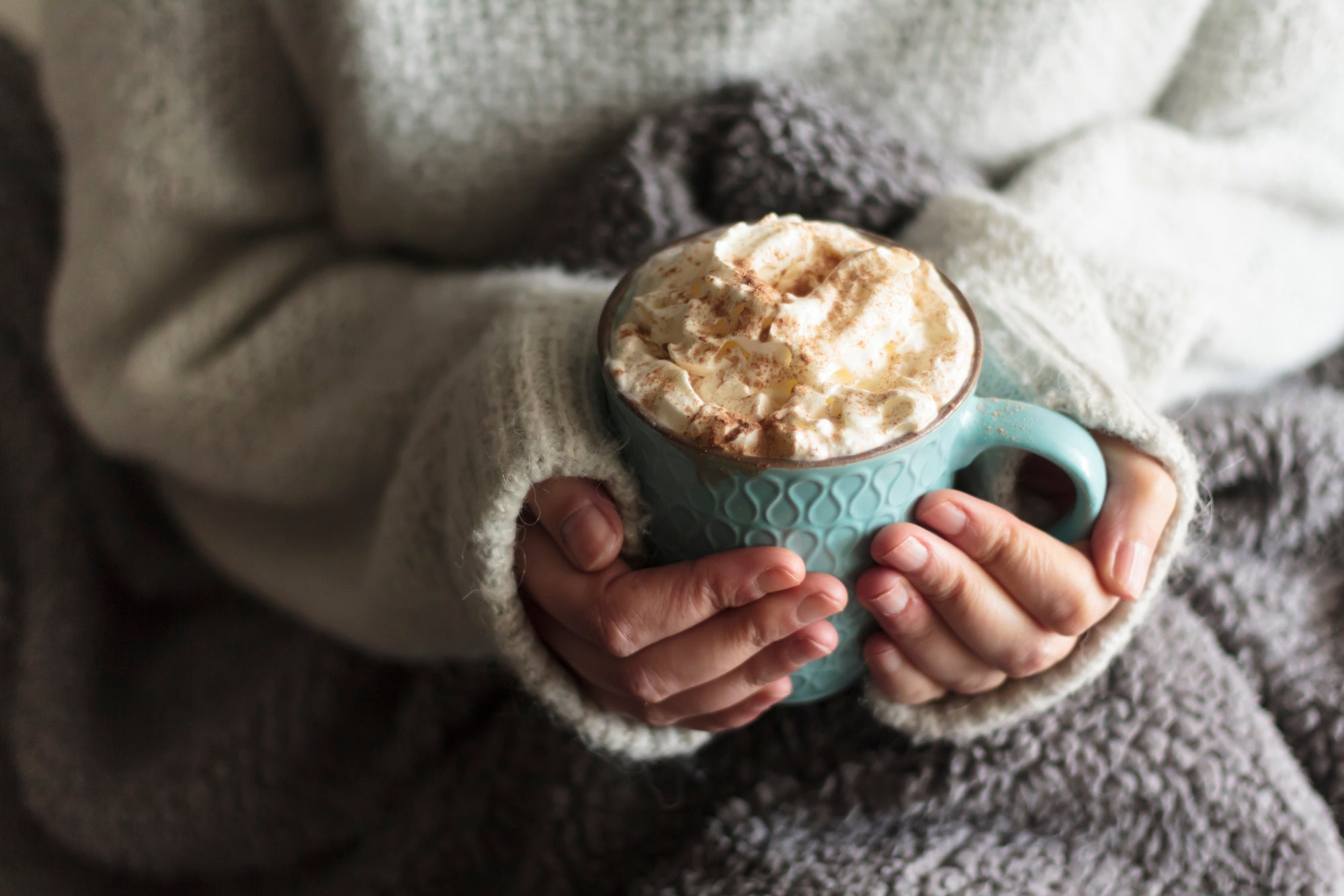 Woman holding cozy cup of cocoa in white sweater and grey blanket