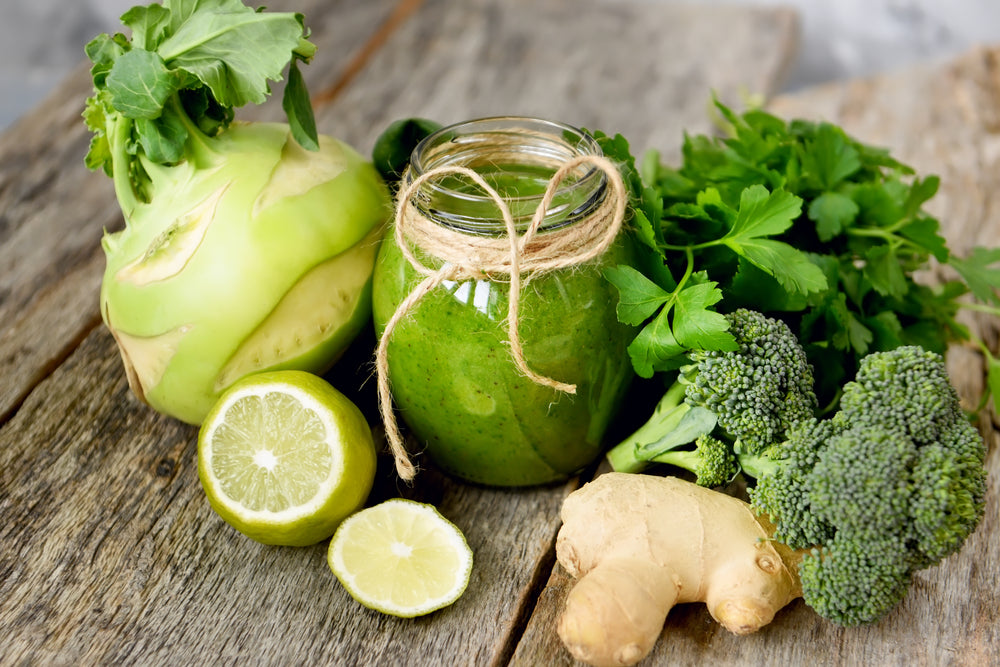 a jar of luminous green detox smoothie wrapped with ribbon string on the neck of the glass surrounded by cilantro, broccoli, ginger and cut lemon on the wooden table.