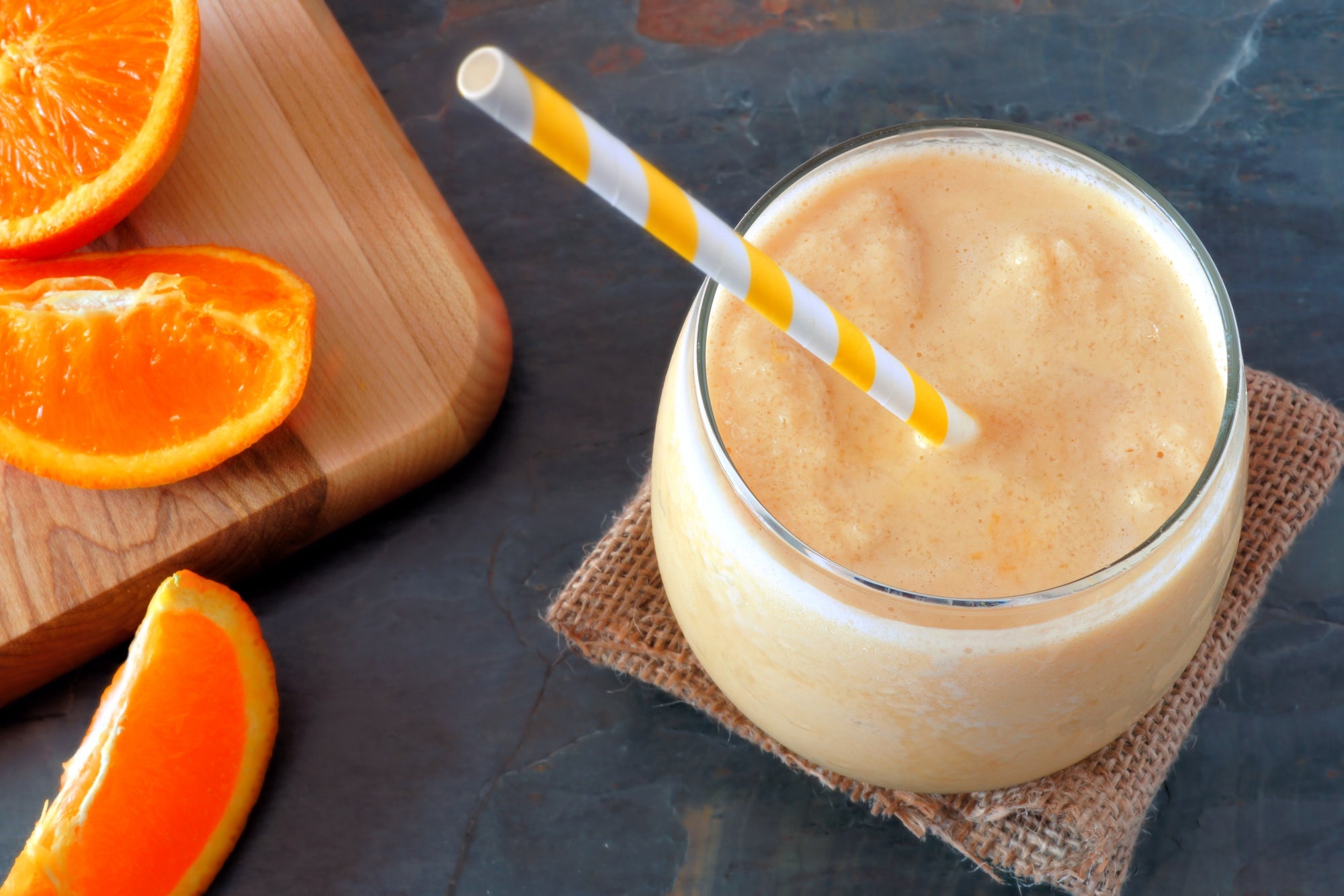 top view shot of a glass of Orange Julius Immunity with a straw, beside a wooden cutting with 2 oranges slices on top and 1 on the surface. 