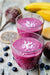 2 glasses of Donna Marie's Energy Proteini frozen berry smoothie garnished with coconut shreds. On the wooden surface are 2 bananas, blueberries, beet root, and a slice of dragonfruit.