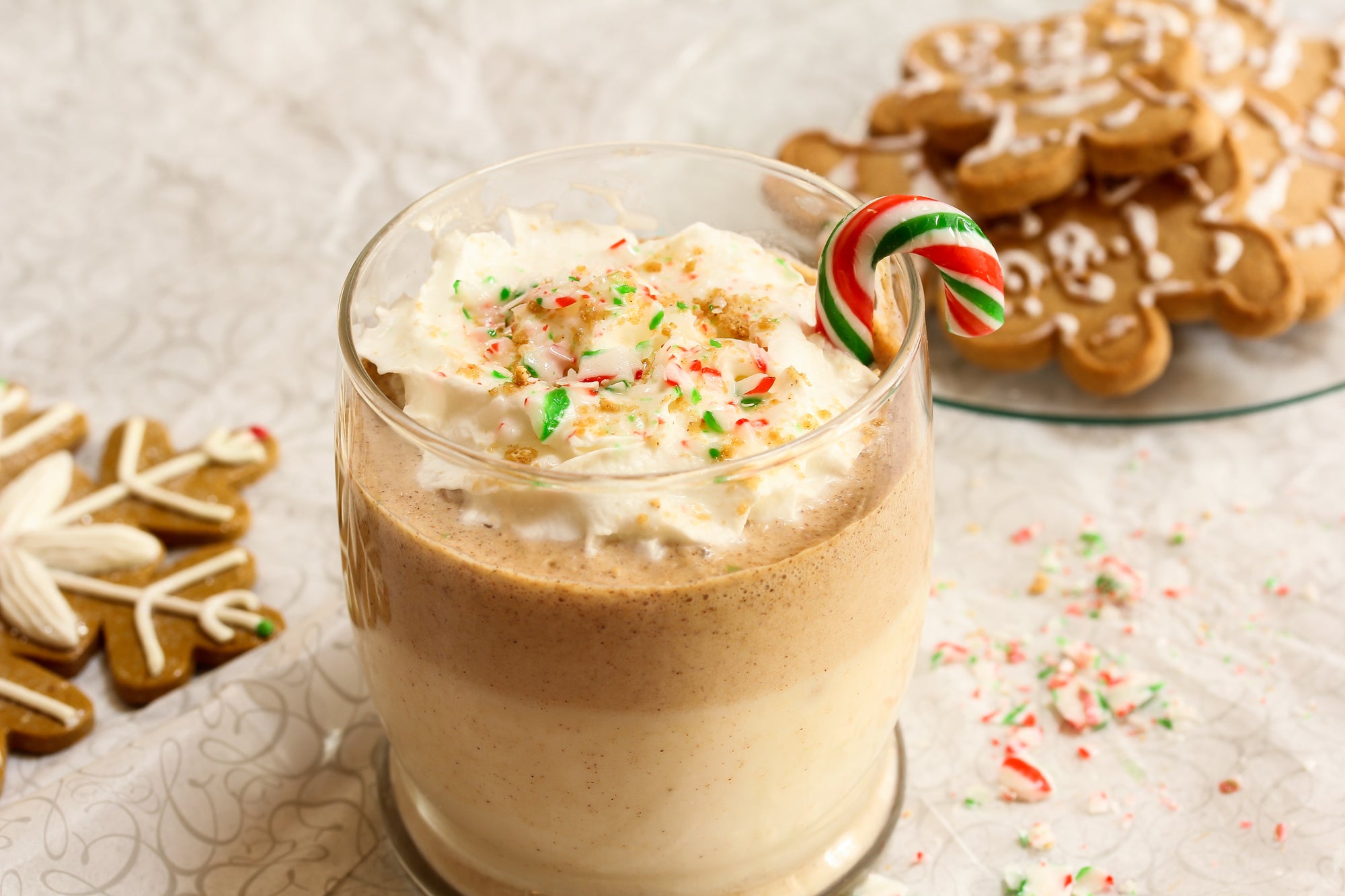 a glass of a gingerbread proteini smoothie topped with whip cream with a transparent plate on the side with gingerbread on top of a white patterned table cloth