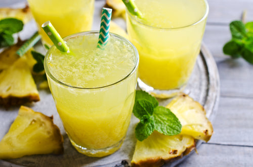 three glasses of pineapple ginger placed on a wooden tray with two green paper straw and a blue patterned paper straw surrounded by slices of pineapple and mint leaves