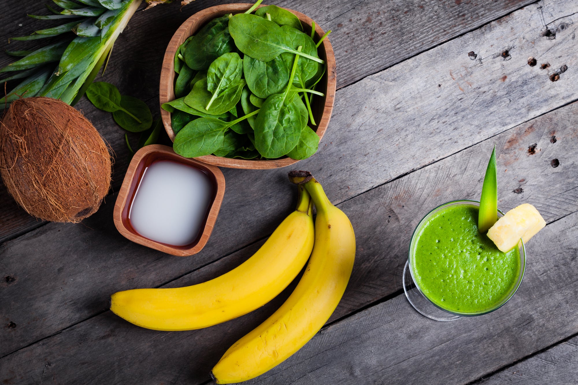 top view shot of 2 bananas, a glass of green smoothie, a bowl of spinach, 1 coconut, 1 small bowl of coconut milk, and pineapple leaf on a wooden surface.