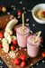 2 glasses of Berry Banana Proteini with straw sprinkled with oats and  surrounded with strawberries, oats, and slices of banana on a wooden cutting board. 
