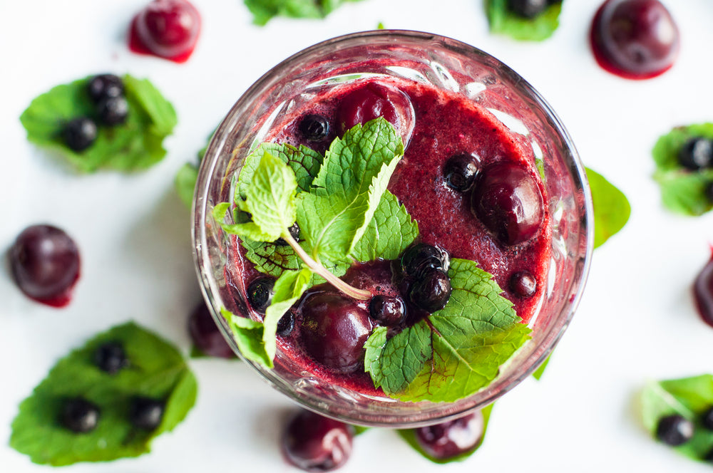 A glass of Cherry Blueberry Smoothie with mint, blueberries, and cherries surrounded by blueberries and cherries