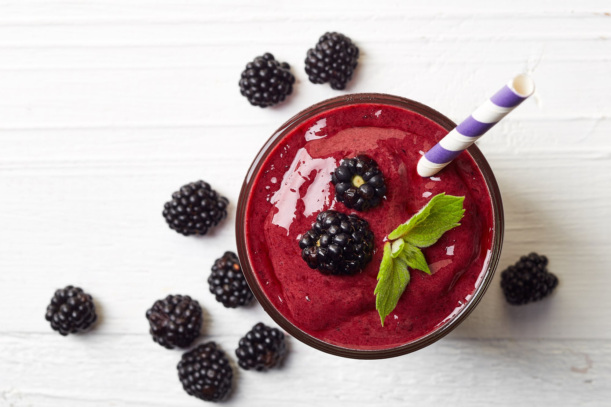 A glass of blackberry smoothie with a straw and mint leaf next to 9 blackberries