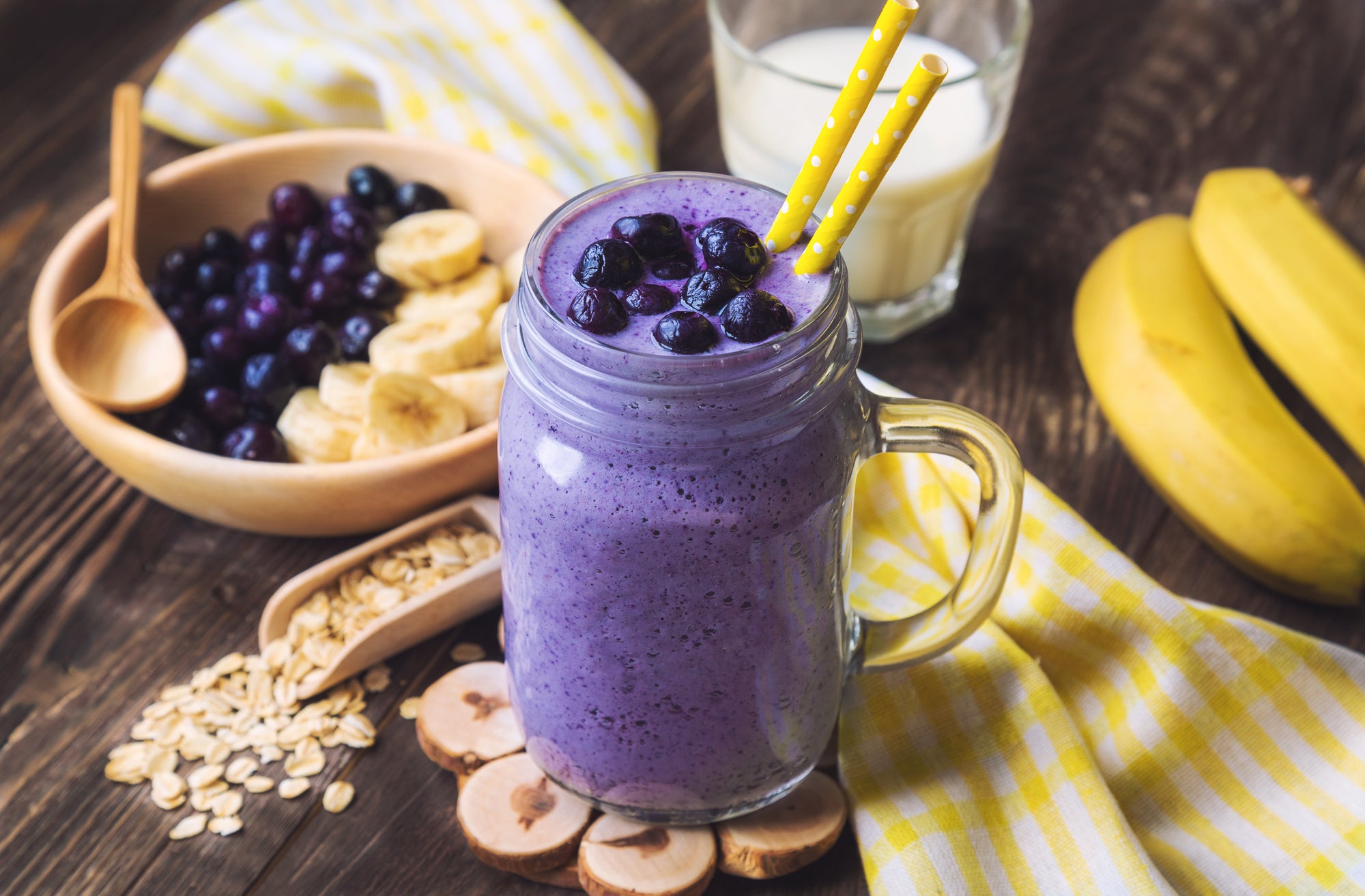 1 jar of blueberry smoothie topped with blueberries with 2 yellow straws behind is a wooden bowl of blueberries with slices of bananas and a wooden spoon beside a glass half full of milk beside 2 bananas and a yellow and white checkered cloth.