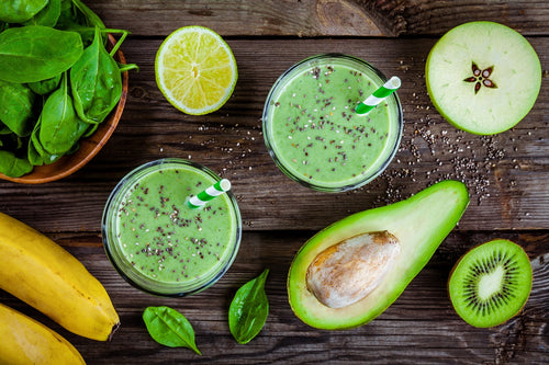 top view shot of 2 glasses of green smoothie sprinkled with chia seeds with a green striped straw surrounded by half avocado with seed, half a kiwi, 1 slice of apple, 1 slice of lemon, 1 bowl of spinach, and 2 bananas on a wooden surface.