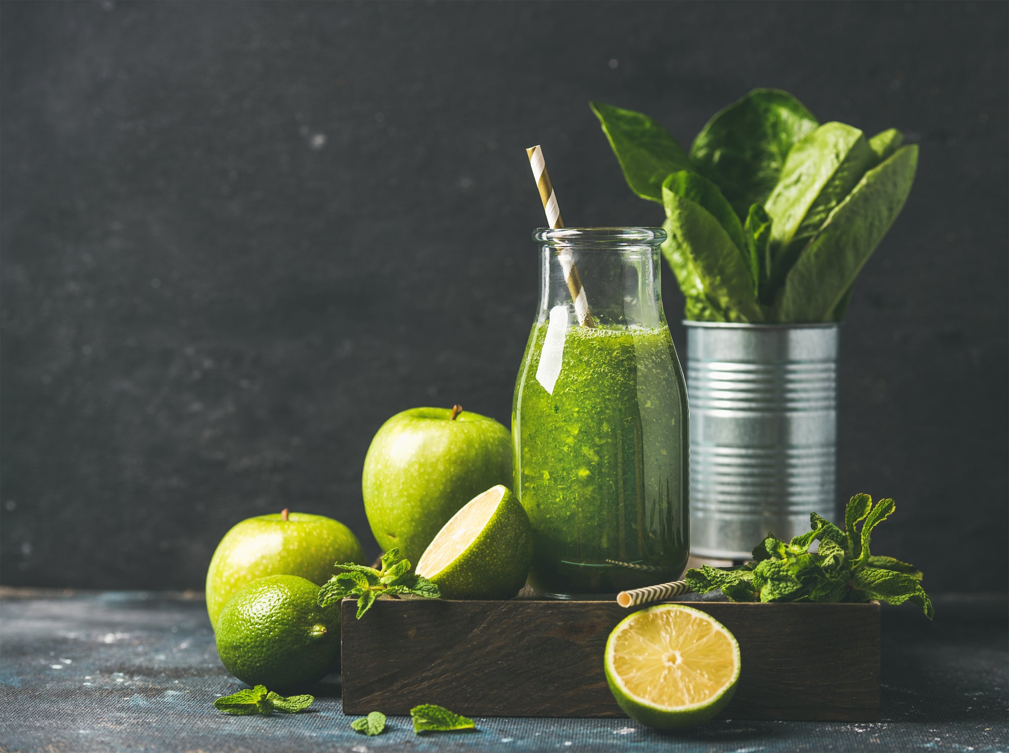 a bottle of citrus pressed greens smoothie - fat burner, a can on the side filled with spinach, one green apple and a half-slice lemon surrounded by mint leaves, a green apple, a lime, and a slice of lime on top of a black-painted wooden table