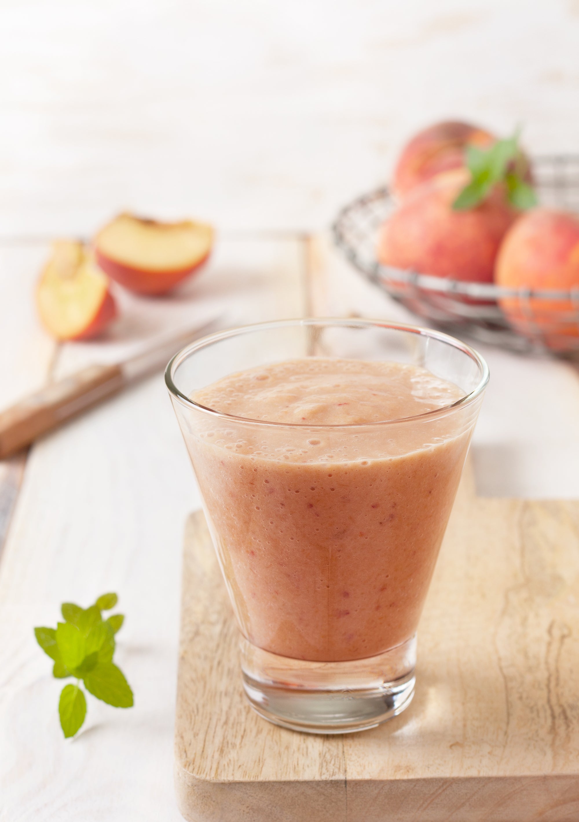 a glass of mixed banana, strawberries, and peaches smoothie on top of a wooden chopping board with knife and slices of two peaches and a fruit basket of peaches