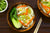 top view of three bowls with two wooden bowls of veggie noodles with spicy miso soup and one small white ceramic bowl of slices of lemongrass and chopsticks on the side on top of a brown wooden table