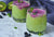side view of two glasses of pressed greens acai smoothie topped with mint with blue-colored cloth and half-cut kiwi surrounded by blueberries and mint on top of a marble table 