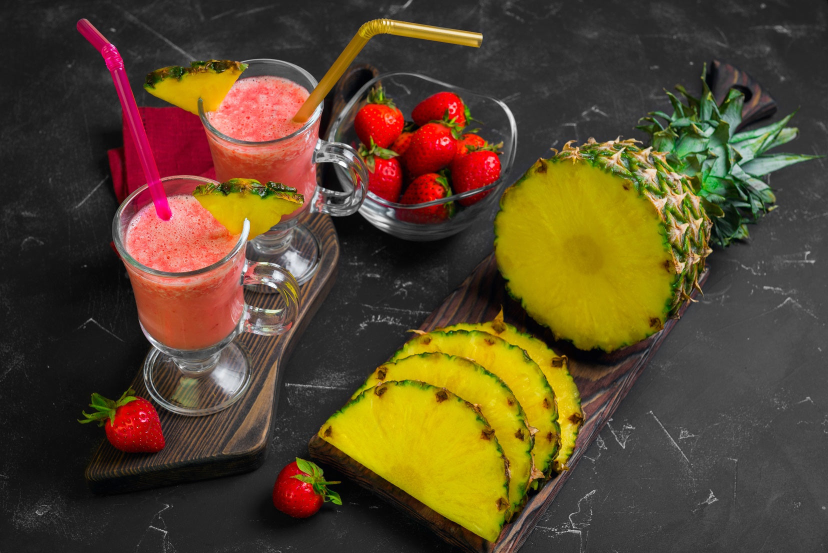 two glasses of signature pineapple chia cleanse smoothie with red and yellow plastic straw topped with sliced pineapple on top of a plank and a sliced on pineapple and a half pineapple on top of a plank and a bowl of strawberry on the side
