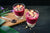 two glasses of pineapple chia acai berry smoothie topped with berries one is on top of a wooden plank with a spoon filled with black powder on the side on top of a black-painted table