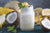 a jar of pina colada smoothie topped with slices of ripe banana with mint and coconut surrounded by  coconut, slices of pears, wedges of lemon, a slice of melon, and a banana with the scissor on the side with mint leaves on top on the top table 
