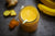 one jar of anti-Inflammatory mango smoothie surrounded by a spoon full of turmeric powder, two slices of ginger, and two pieces of bananas on top of the table