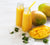 two bottles of mango greens smoothie with yellow plastic straw surrounded by two mangoes, a sliced mango cut in cubes, and mint leaves on top of a white table