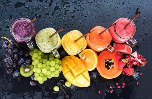 top view of mason jars with different smoothie flavors and a brown paper straw surrounded by blueberry, grapes, a sliced of kiwi, sliced of mango and orange, a sliced of papaya and strawberries, a sliced of watermelon, and a cut of pomegranate