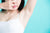 Woman in white tank top on a baby blue background holds her nose as she extends her arm upwards 