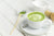 a cup of matcha greens latte on top of a saucer with a teaspoon on the side with a small bowl of matcha powder with a small bamboo spoon and an extra empty cup on the side