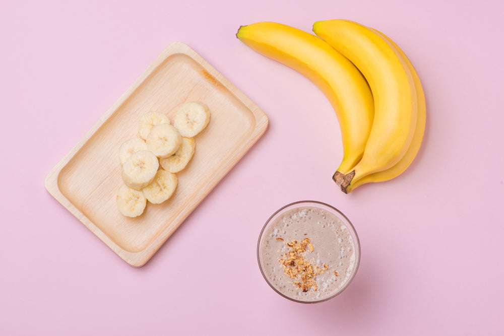 a glass of Banana Nut Proteini with crushed nuts next to 2 pieces of banana and sliced bananas