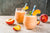 two glasses of peaches 'n' cream smoothie with blue and white patterned paper straw and a sliced of peach and mint on top surrounded by one whole peach and slices of peaches