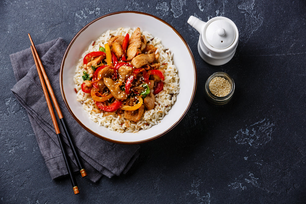 top view of Rice Bowl topped with sauteed vegetables with slices of chicken on top of a blue folded table with chopsticks and a small jar with sesame seeds and a white cup with a lid on top of a blue-painted concrete table