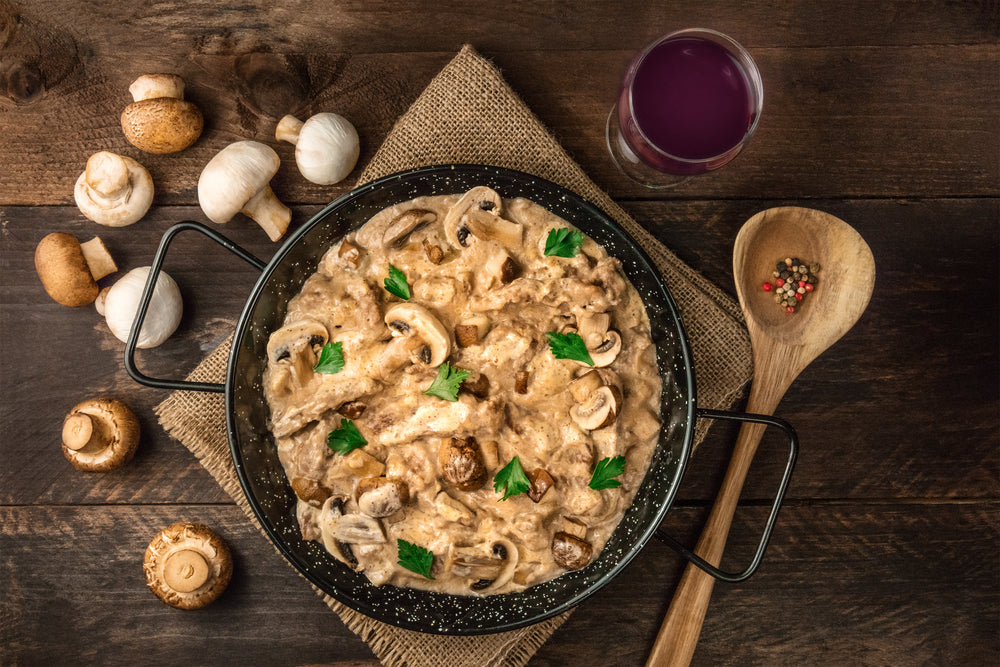 a top view of skillet full of mushroom stroganoff on top of a weaved clothe with whole mushrooms on the side and a wooden ladle of peppercorn and a glass of grape juice on the side