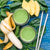 top view of two glasses of pineapple chia cleanse surrounded by spinach and a half-peeled banana and three slices of pineapple and two gray patterned paper straw on top of a blue-painted table