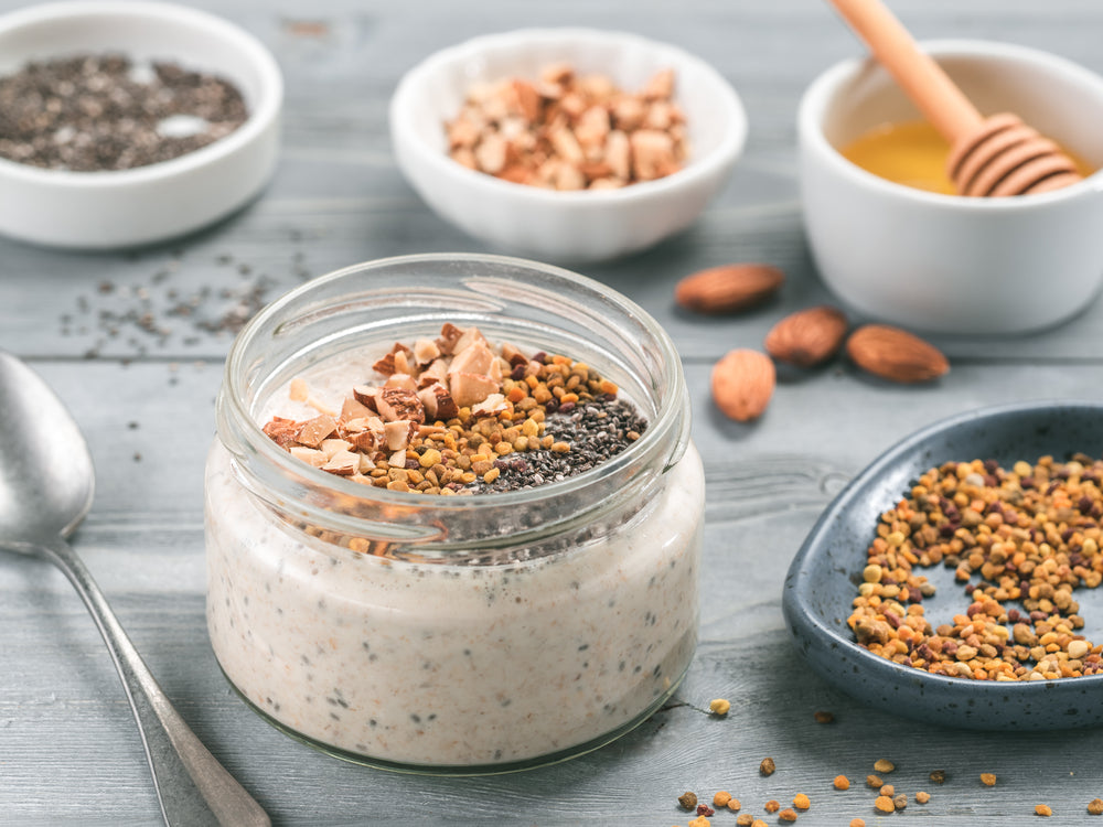 one jar of pudding topped with chia seeds, crushed almond and dried cherries surrounded by a bowl each ingredient, chia seeds, almond, honey, and dried cherries with a spoon and almond nuts on the side on top of a wooden table