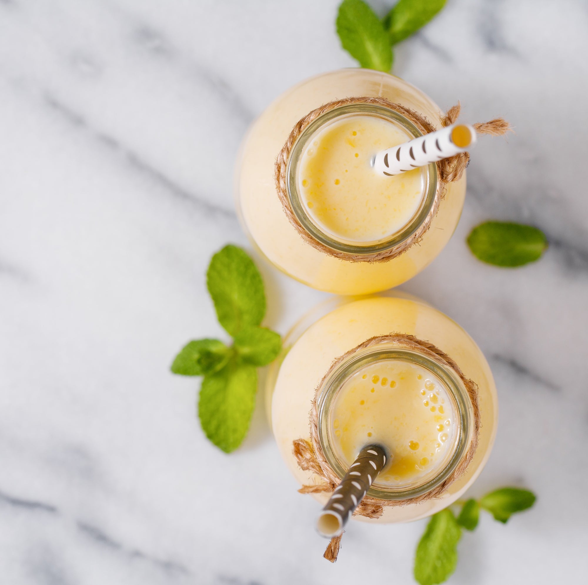 top view of two jars of acerola pineapple smoothie with string on the lid and brown and white polka dot patterned paper straw surrounded by mint leaves on top of a marble table
