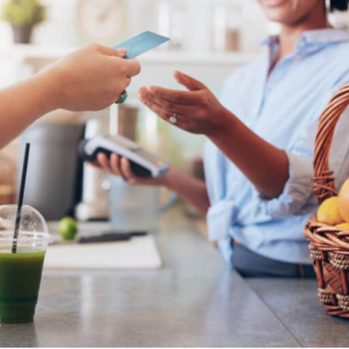 A woman handing a credit card to a woman infront of her wearing a blue long sleeve folded in half and a wooden basket full of fruits and a plastic cup with cold-pressed green juice with blue paper straw on top of a counter table.