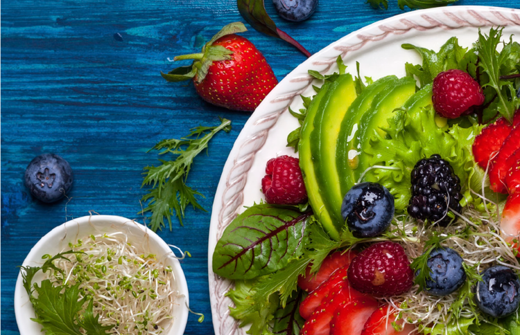 top view shot of a plate of salad with kale, spinach topped with 4 different kinds of berries, avocado slices, and chia sprouts beside a smaller bowl of chia sprouts. On the blue surface is 1 strawberry, 2 blueberries, and some green leaf vegetables.