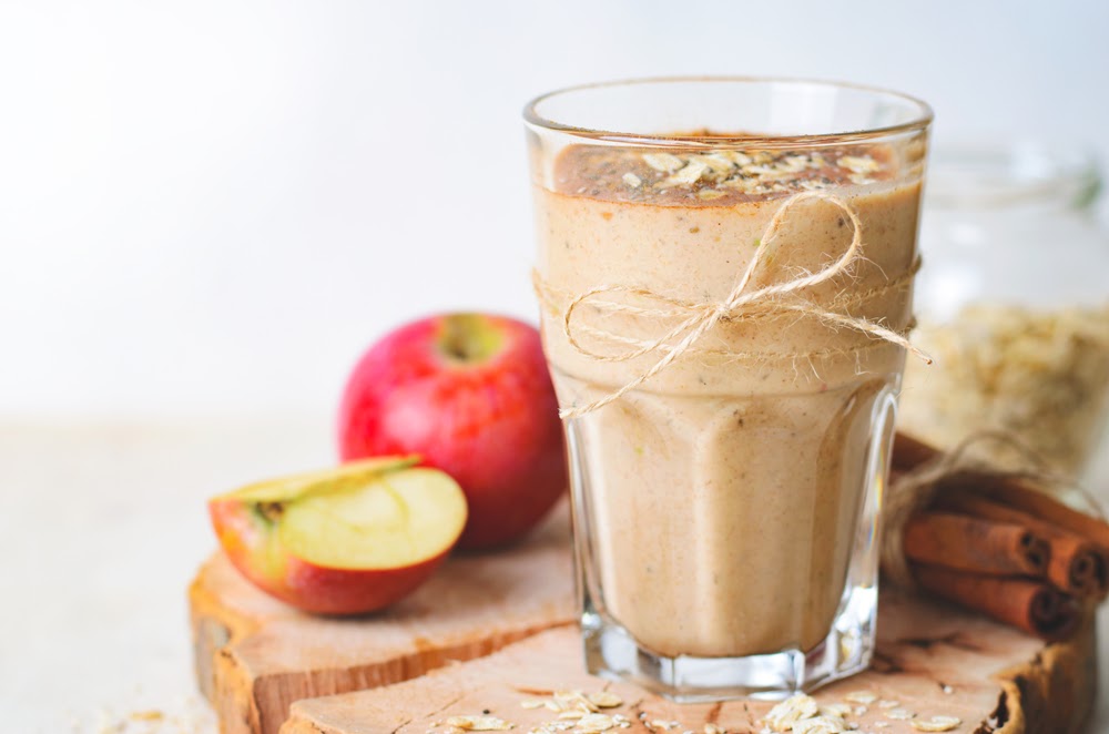 A glass of Apple smoothie wrapped with a twine string. The glass is on top of a wooden board with apple, apple slices, cinnamon sticks, and a jar of brown sugar on the side.