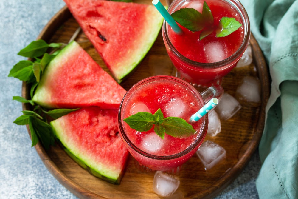 2 red smoothie with ice in a tall glass and pieces of watermelon and ice on top of a wooden tray