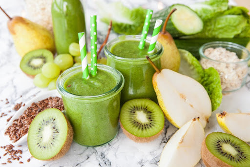 Image of 2 green shakes in a glass with pears and kiwi surrounding it on a white table