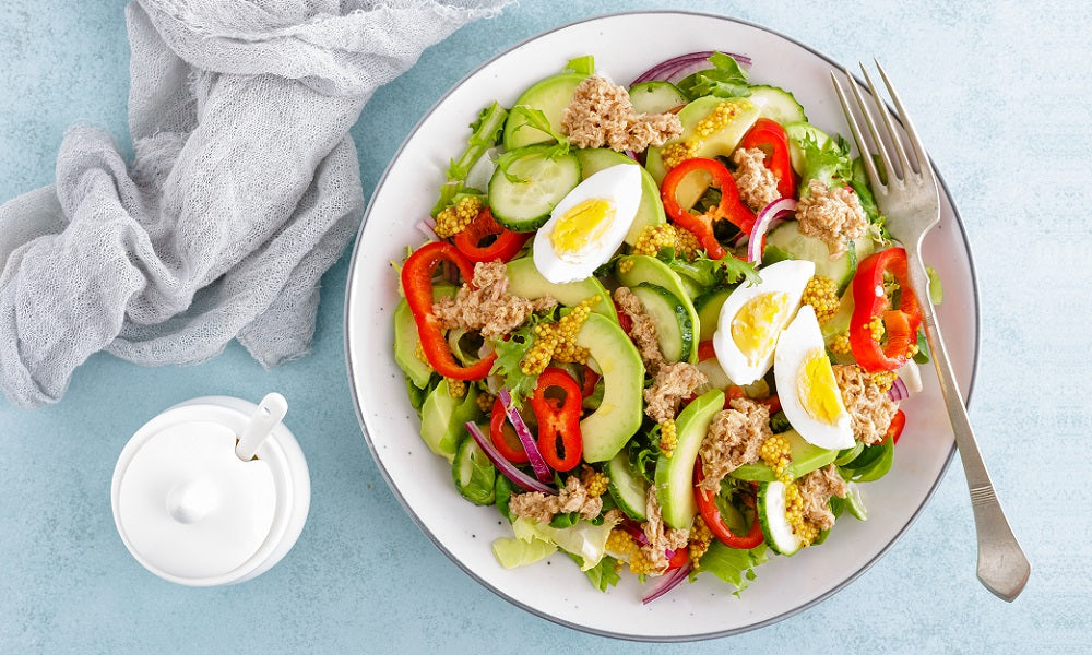 Top view image of a bowl of salad with avocado, red bell pepper, slices of hard-boiled eggs, slices of cucumber, slices of red onion, and tuna flakes. Beside is a white face towel and a white tumbler.  